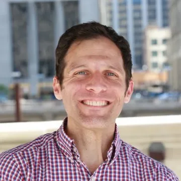 Jonathan R. Davis smiling outdoors in a red checked shirt, with city buildings behind him